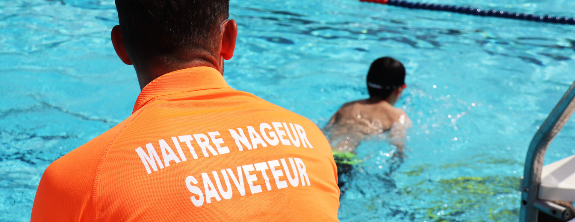 Maitre nageur sauveteur LMV en polo orange sur lequel est écrit MAITRE NAGEUR SAUVETEUR pris de dos en train de surveiller un enfant qui se bagne à la piscine de plein air.